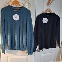 Two sweaters hanging on wooden hangers on a door. The blue sweater marked A and black sweater marked B.