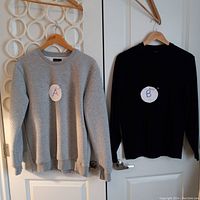 Two medium men's sweaters hanging on wooden hangers on door handles, labeled A and B for identification purposes. Sweater A is grey ribbed, sweater B is black slightly ribbed.