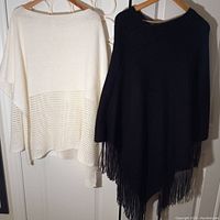 White acrylic shawl and black fringe shawl hanging on wooden hangers side by side against white door, showing full front view of both shawls.