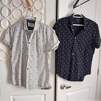 Photo of two short-sleeved shirts hanging on door handles, one white with black stripes and pattern by Jack & Jones and one dark blue with white specks by Zara Man.