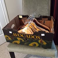 Box labeled 'Avocados' contains 20 wooden clothes hangers with metal hooks in two different wood finishes inside resting on a chair.