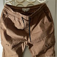 Close-up of front view of brown Urban Man jogger pants showing elastic waistband with black drawstring, side pockets, and cargo pockets on the legs.