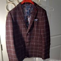 Front view of brownish maroon dress jacket with light blue plaid pattern hanging on a red hanger showing paisley patterned lining and chest pocket with matching pocket square.