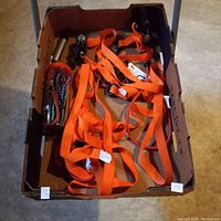 Box containing two orange ratchet tie down straps with metal ratchet mechanisms and several bungee cords with hooks.