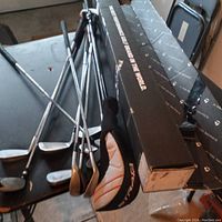 Photo showing 10 golf clubs including irons, wood/driver with TaylorMade cover, and putter on a black table near boxes.