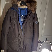 Front view of a brown heavy winter jacket with multiple pockets, zipper and snap closure, hanging on a door handle.