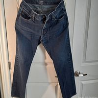 Single pair of men's blue denim jeans hanging on a hanger in front of a white door, showing full front side.