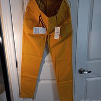 Full view of mustard-colored pants hung on a door, showing front side, button closure, and attached tags.