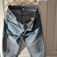 Light blue denim skinny jeans with ribbed thigh and knee panels, hanging against a door, showing front button zipper and pockets.