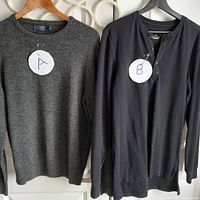 Pair of sweaters hung on hangers against wall, labeled A and B for identification. Sweater A is dark grey J.Crew wool crew neck, sweater B is black lightweight henley shirt.