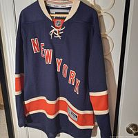 Front side of dark navy blue New York Reebok hockey jersey showing 'NEW YORK' in white and orange lettering with stripe detailing and lace-up neckline.