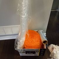 Photo showing a large orange roll of bubble wrap, clear plastic wrap roll, and loose foam packing peanuts in a box next to a dog.