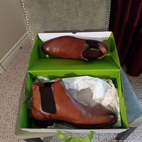 Pair of men's brown leather Chelsea boots by PEGABO inside green shoebox, showing left shoe on box base and right shoe resting on top with white tissue paper.