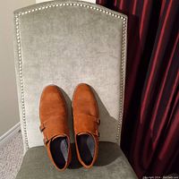 Top-down front view of tan suede men's shoes on gray upholstered chair, showing monk strap buckle detail and rounded toe.
