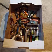Top view of an open box showing a large assortment of household tools including pliers, screwdrivers, chisels, hacksaw, and hardware items. Tools show signs of light use.