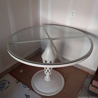 Full view of the round glass table with white metal pedestal base from the side corner showing overall structure and size.