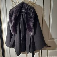 Full view of the black wool jacket hanging on a door with the removable dark fur collar visible around the neckline and draped over the shoulders.