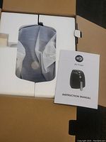 Photo showing the Simply Ming air fryer nestled in white foam packaging with a protective white mesh wrapping and the instruction manual beside it.
