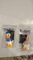 Two Lego Disney minifigures, Donald Duck and Daisy Duck, sealed in plastic bags each with a black baseplate behind them.