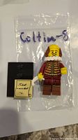 Photo showing the William Shakespeare LEGO minifigure standing on a black base with a cream-colored scroll accessory, posed in front of a labeled plastic bag.