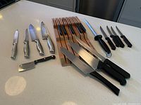 Overview of all knives including large chef knives with metallic handles, cleaver, small black handled knives, knife block with steak knives, sharpening rod, and other cutting utensils on kitchen counter.