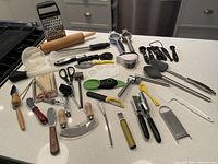 Overview of all assorted kitchen accessories on a white countertop including graters, cutters, rolling pin, peelers, garlic press and others
