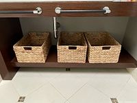 Three rectangular woven storage baskets placed side by side on an open shelf under a sink.