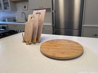 Three sizes of bamboo cutting boards standing upright on a small wooden stand and a circular bamboo lazy susan placed next to them on a countertop.