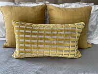 Three gold-tone bedroom accent pillows arranged on a bed; two square pillows in back and one rectangular patterned pillow in front