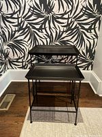 Set of two black metal nesting tables against a patterned black and white wall, showing their relative sizes and tray tops.