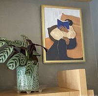 Framed painting hung on a wall above a wooden shelf with a glass planter containing leaves.