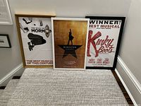 Three framed Broadway theater posters for Hamilton, Kinky Boots, and The Book of Mormon arranged on a carpeted floor against a wall.