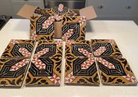 Box of sixteen decorative ceramic tiles with intricate geometric and floral patterns in black, gold, rust, and red tones arranged in front of box.