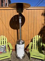 Full view of the patio heater standing outdoors between green chairs against a wooden fence, showing overall condition and size.