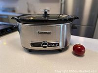 KitchenAid 6-quart slow cooker with stainless steel finish and glass lid on kitchen countertop with red apple for size reference.