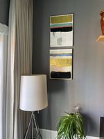 Two large abstract paintings displayed vertically on a wall above a floor lamp and plant. One portrays bold black, coral, greenish, and gold horizontal bands with white background.