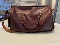 Front view of smooth brown leather duffel bag with handles and shoulder strap laid on surface.