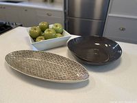 Three serving bowls on a kitchen counter: brown Crate & Barrel round bowl, Wonkiwear oblong patterned bowl, and white Crate & Barrel square bowl with artificial green apples inside.