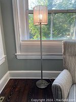 Photo of a skinny standing reading lamp with silver metal finish and beige cloth shade placed by a window next to a gray upholstered chair.