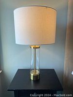 Single lamp on black table showing acrylic clear tall cylindrical base with brass metal top and bottom and linen drum shade