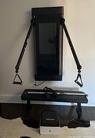 Wide shot of Tonal wall unit mounted on white wall with adjustable arms and paired with padded bench; accessories and Theragun in foreground