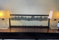 Framed black and white print depicting row of trees on a console table between two lamps