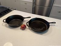 Photo of two cast iron skillets side by side on a kitchen counter with an apple for size reference.