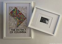 Two framed prints placed flat on table: colorful DC neighborhood map and small black-and-white Snowmaggedon photo.