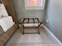 Wood luggage stand positioned near a beige upholstered bed on a carpeted floor, under a small window showing exterior brick.