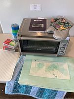 Front and top view of Breville smart toaster oven showing manual and accessories on top and beside it including pot holders, plastic cups, matchboxes, drying rack, placemats, and runner.
