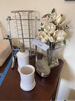 Photo showing silver toilet paper holder, silver hand towel holder, artificial cream and white flowers in clear glass vase, white ceramic soap/lotion dispenser and tumbler, silver soap dish with lid placed on wooden surface.