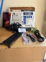 Korg D1200 digital recording board with foot pedal, various cables and manuals arranged on a cardboard surface with packaging box behind.