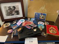 Wide view of Disney collectibles on table including pins, medallions, a black pouch, red ornament, a framed photo, and 50th anniversary ticket folder