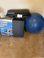 Photo showing black workout bench, PRI folding fitness mat in packaging, and a large blue yoga ball on carpet